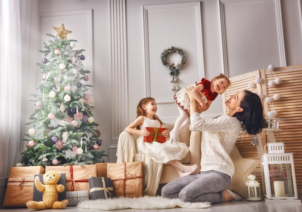 mother holding a little girl laughing, another little girl sitting with a present in her hand, and a Christmas tree with presents underneath