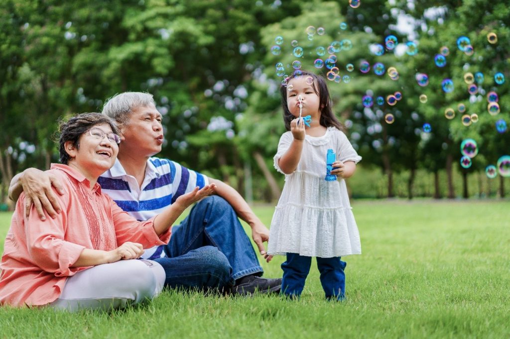 little girl blowing bubbles, and elderly couple sitting on the grass laughing