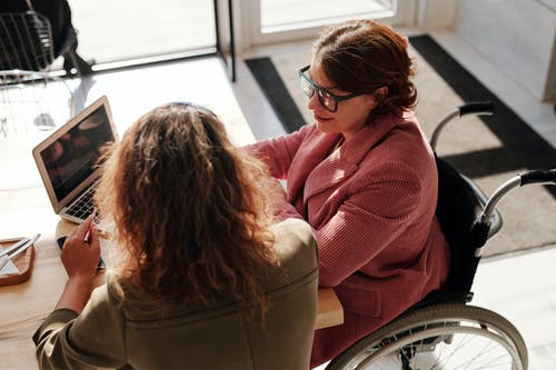 two women, one of them in a wheelchair, using a laptop and talking