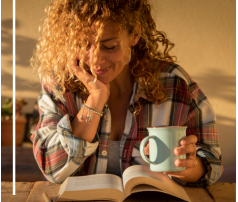 woman smiling and looking at a book with her chin resting in hand and coffee mug in her other hand