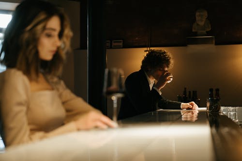 slightly blurred woman in foreground sitting at bar with man wearing suit in background sitting at the other end of the bar