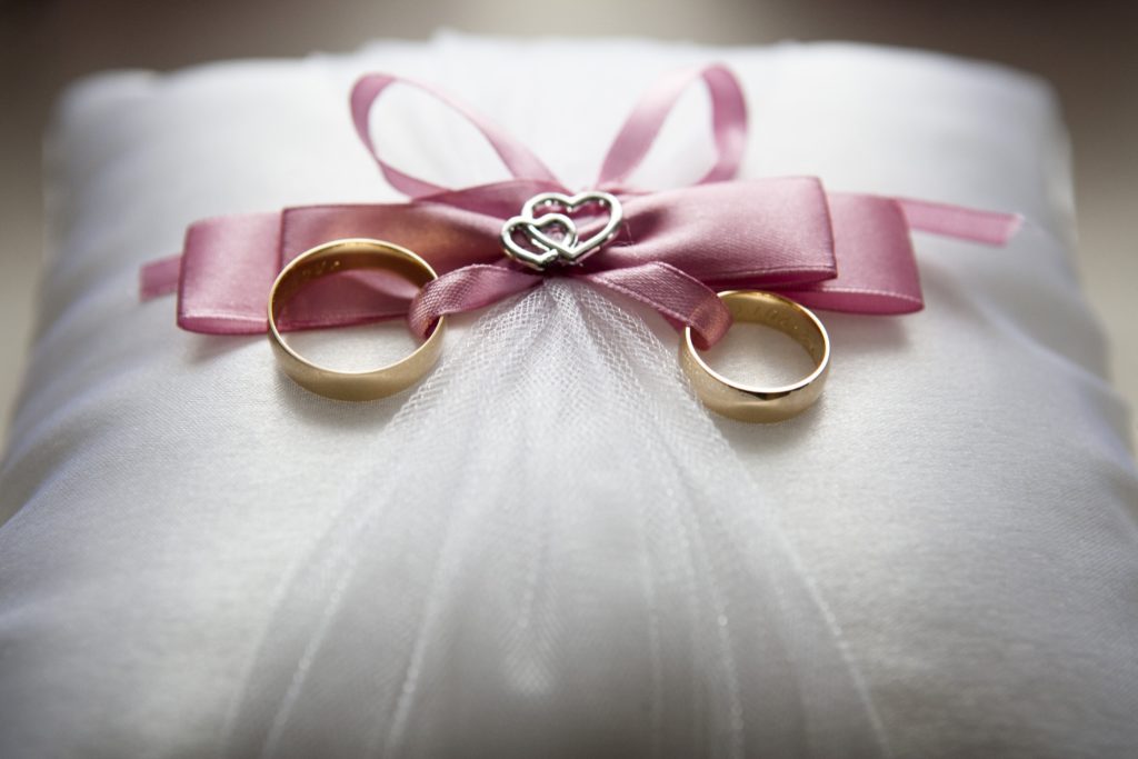 two gold wedding rings resting on a white pillow with a pink ribbon tied into a bow